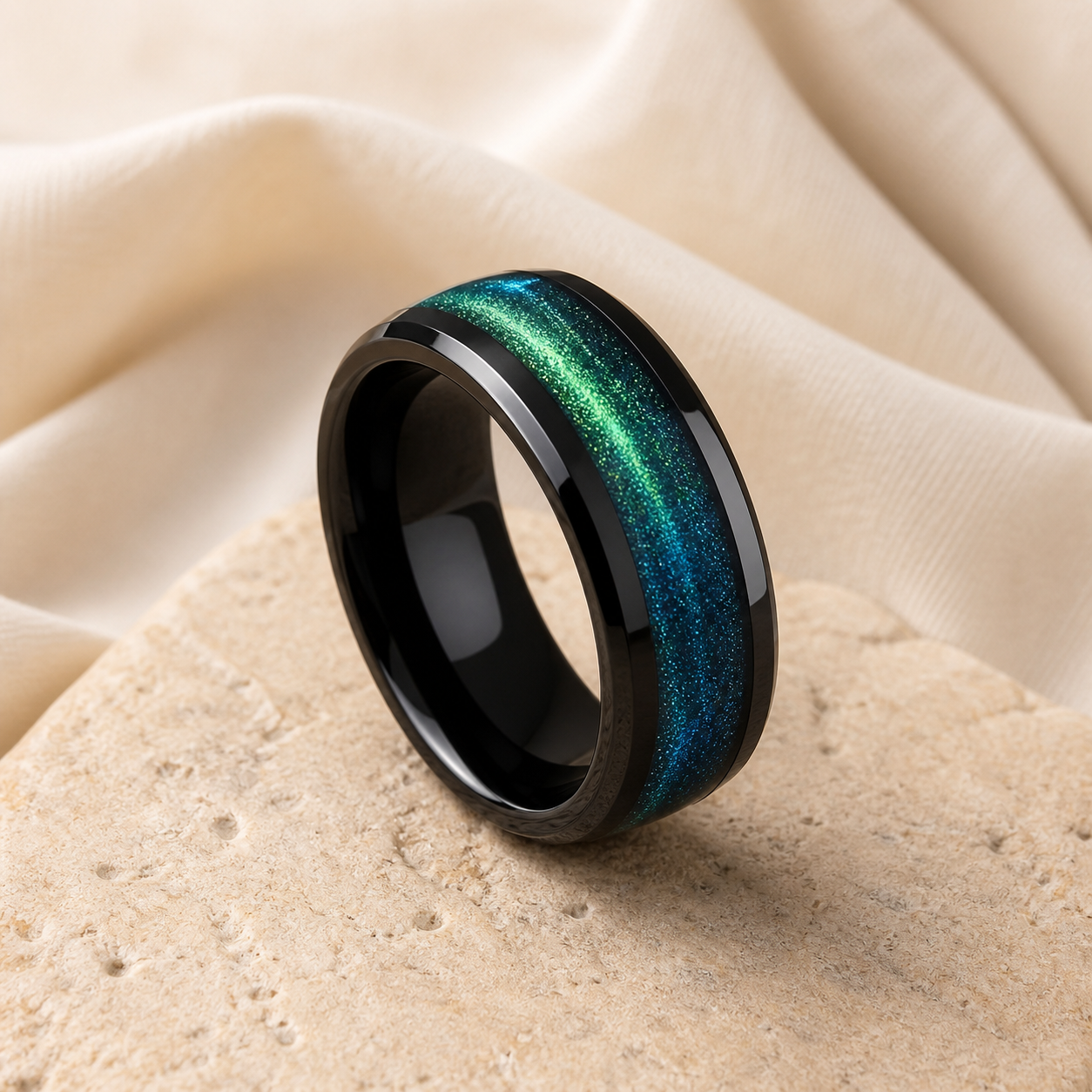 Black tungsten ring with a green and blue aurora-style inlay displayed on warm beige stone