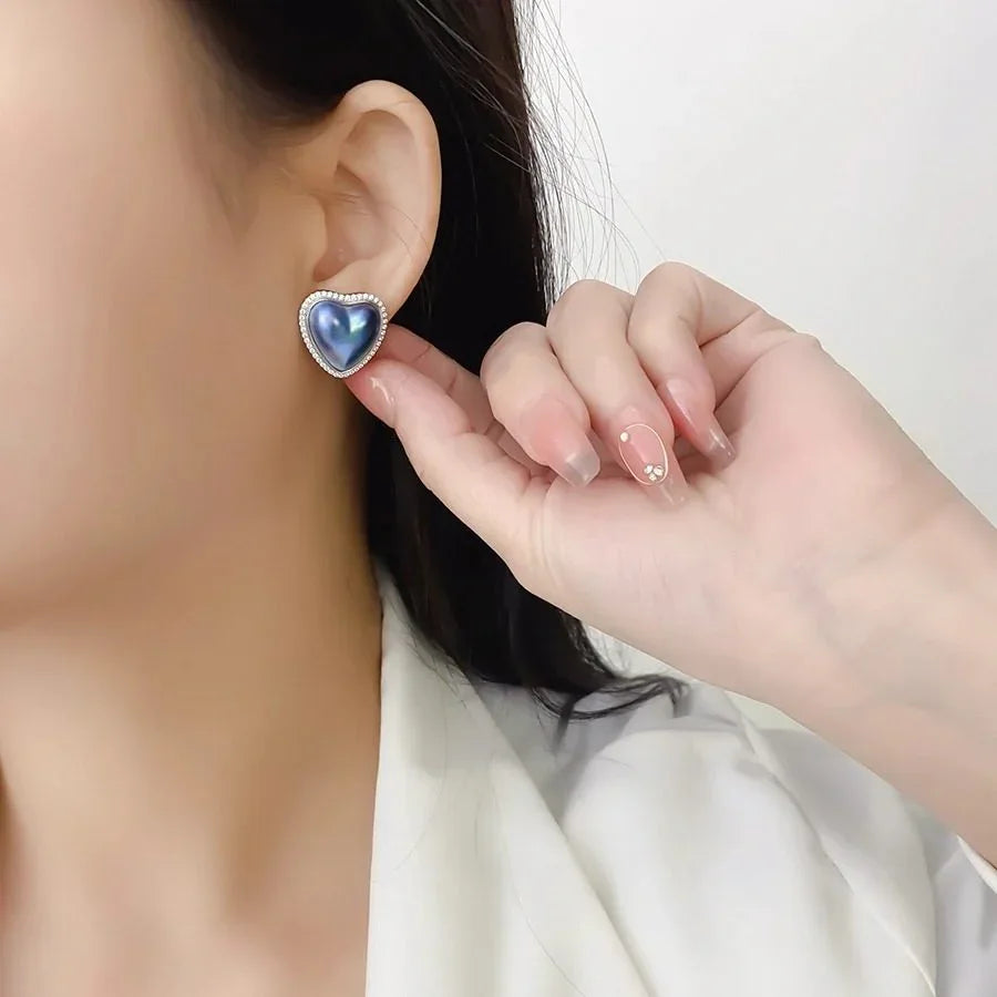 Blue Iridescent Pearl Heart Halo Stud Earrings — left-ear worn close-up with a fingertip indicating scale