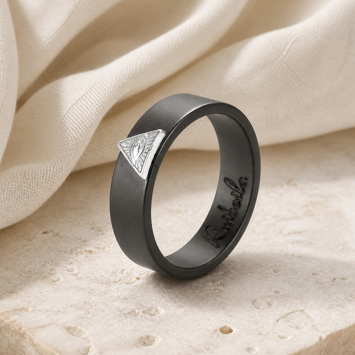 Charcoal ceramic ring with a raised silver-tone triangular eye motif on warm beige stone