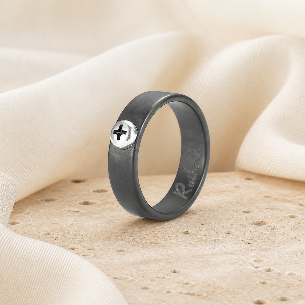 Charcoal ceramic ring with a raised silver-tone cross screw displayed on warm beige stone
