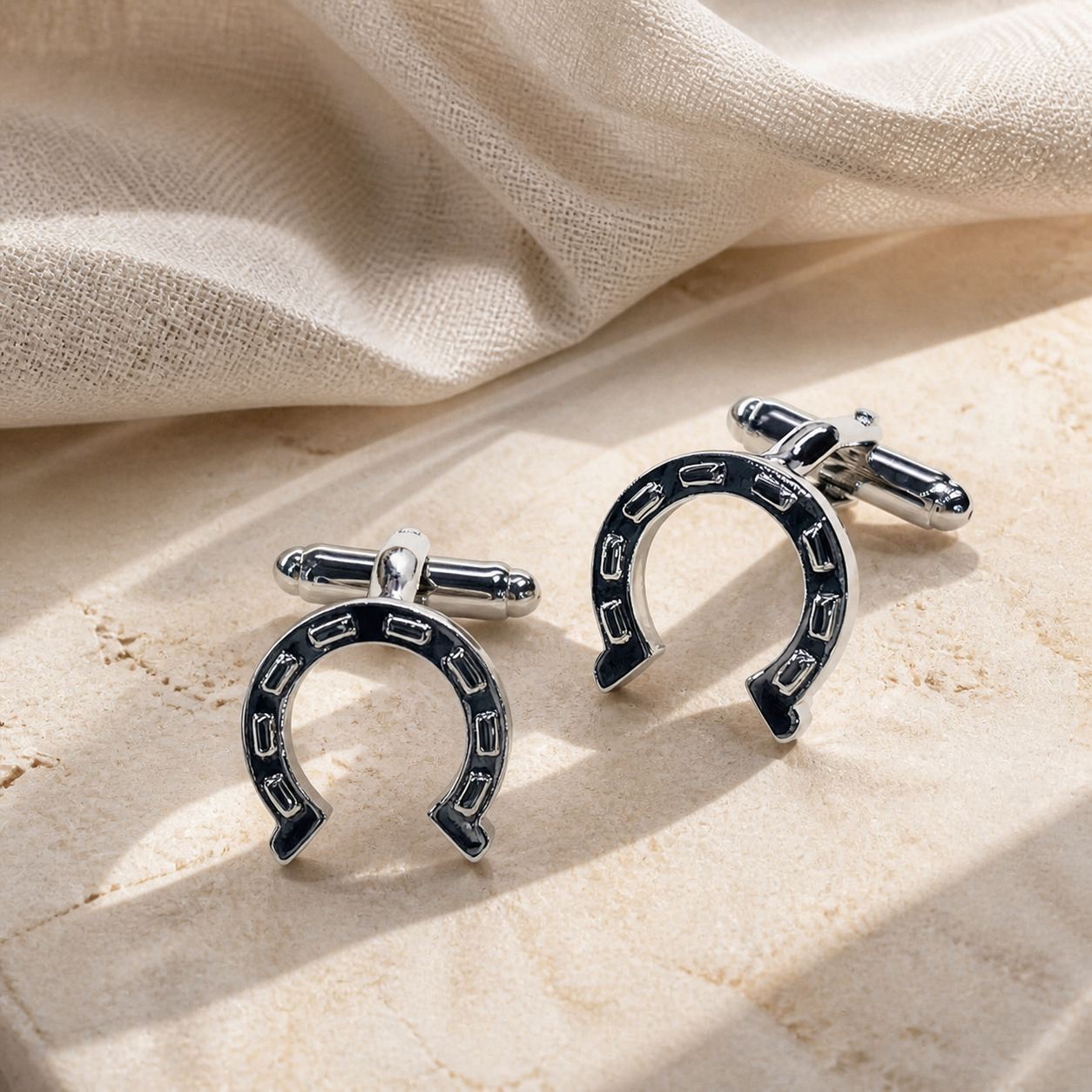 Pair of silver-tone horseshoe cufflinks with dark enamel faces displayed on warm beige stone