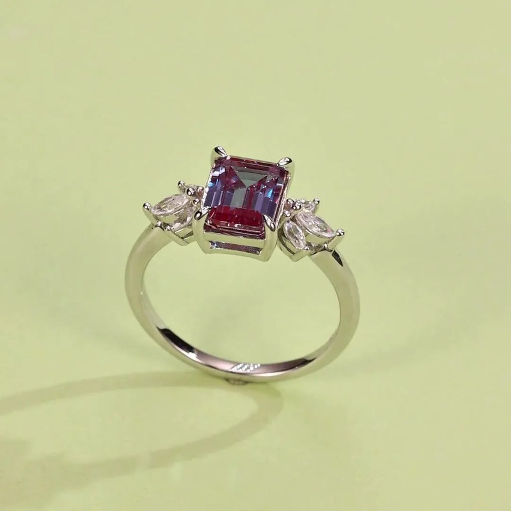 Emerald-cut Alexandrite open ring with leaf side accents, angled product view on a colored background