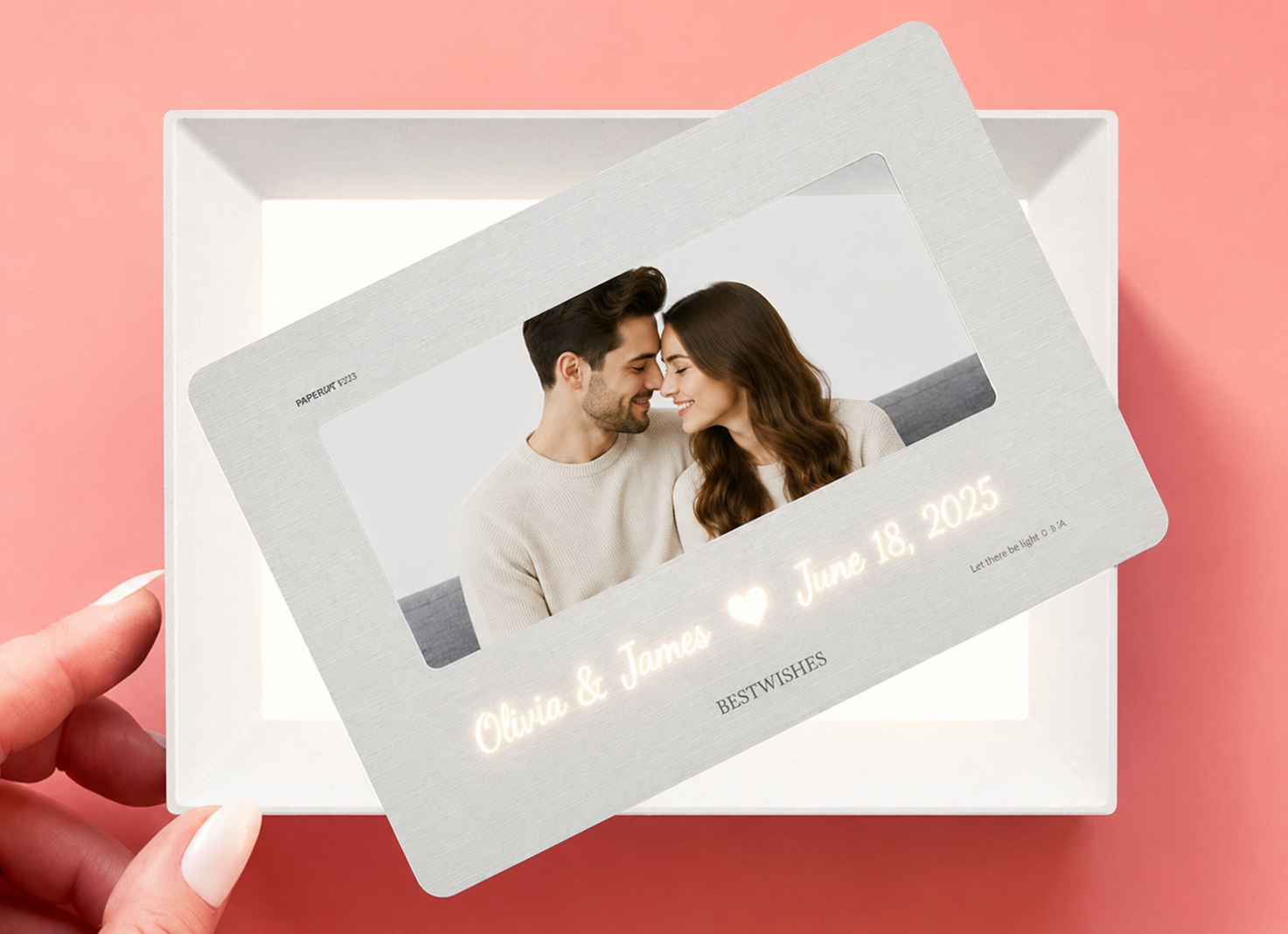 Hand holding personalized hidden message film card showing translucent paper film card with couple photo and glowing script names Olivia and James heart June 18 2025 in warm LED light above white LED light frame base on pink background