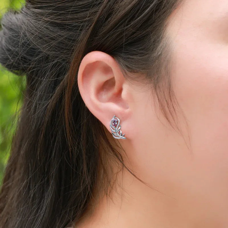 Feather-shaped stud earrings with oval Alexandrites, shown worn on an ear for scale