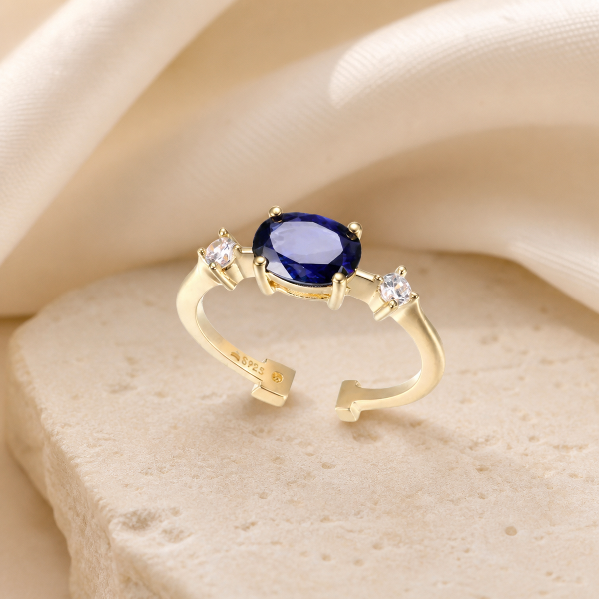 Gold-tone open ring with an oval blue sapphire and two round clear accent stones on beige stone