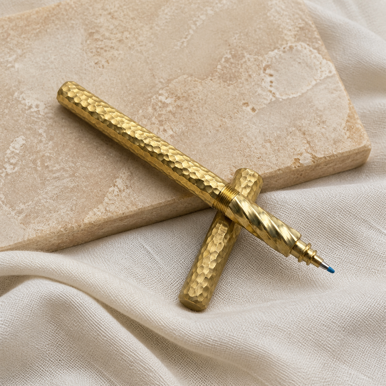 Hammered brass two-in-one pen with its matching cap arranged on beige stone and ivory linen