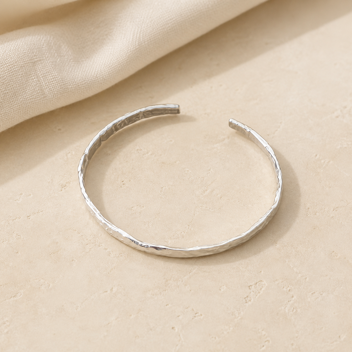 Minimal hammered silver-tone open cuff bracelet on warm beige stone with ivory linen