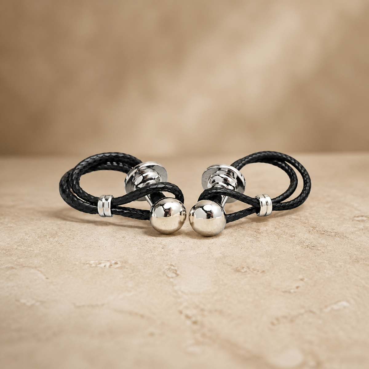 Pair of black braided leather cuff bracelets with polished silver-tone ends on beige stone