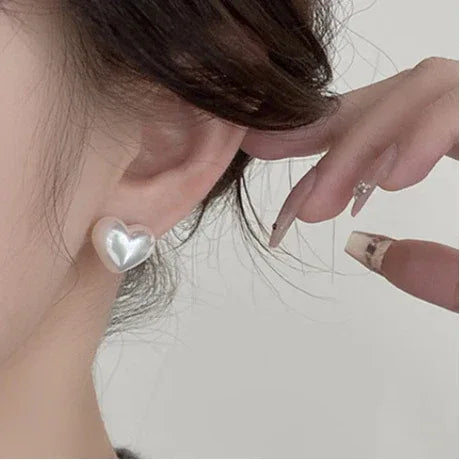Iridescent Pearl Heart Stud Earrings — right-ear worn close-up with a fingertip indicating scale
