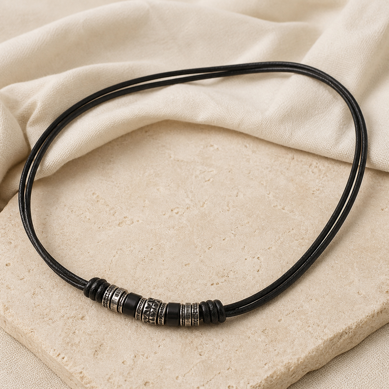 Black double-cord leather necklace with dark and engraved silver-tone beads on warm beige stone