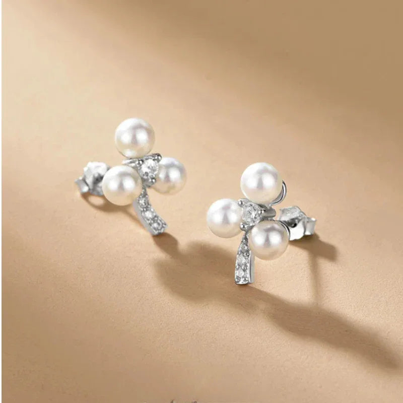 Three-Pearl Bow Stud Earrings in Platinum-Plated S925 Silver — angled pair on a beige surface showing both post backs