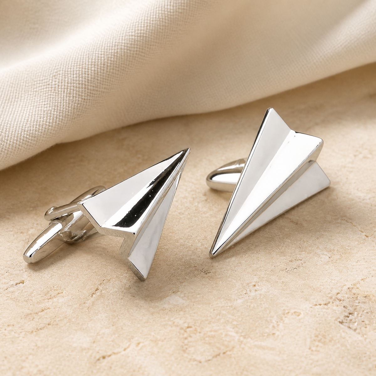 Pair of polished silver paper plane cufflinks displayed on warm beige stone with ivory linen