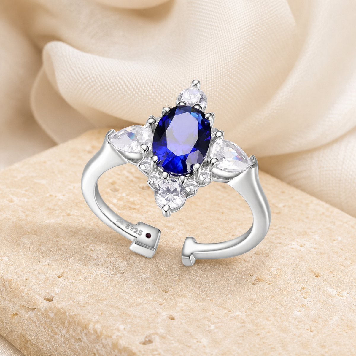 Silver-tone open ring with an oval blue sapphire and clear round and marquise side stones on beige stone