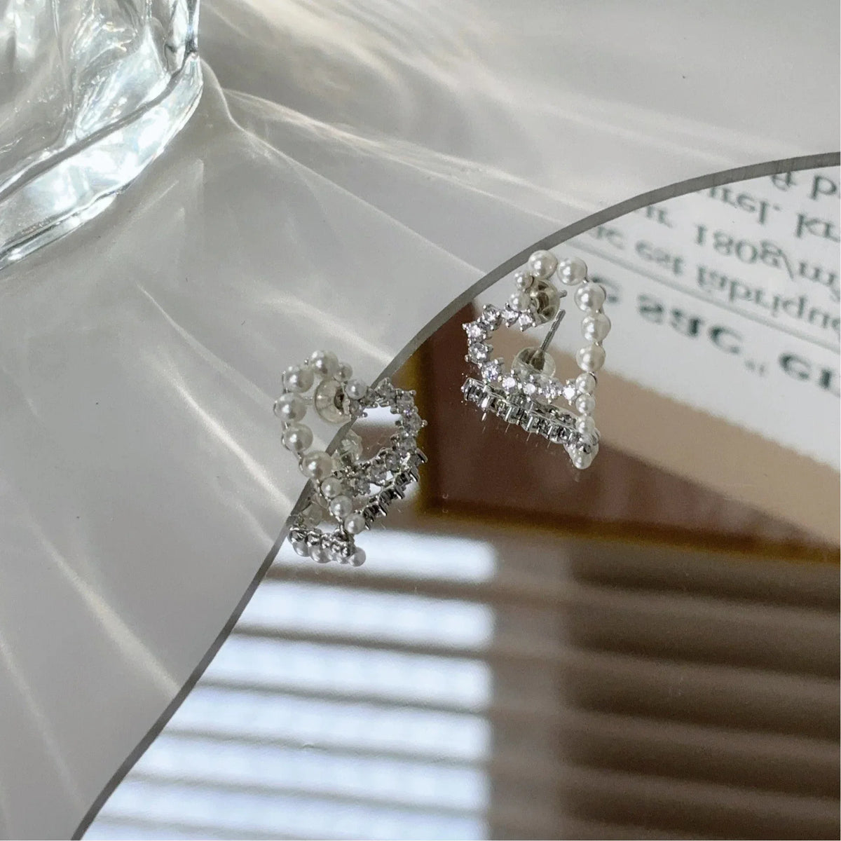 Pearl and Textured Open Heart Stud Earrings — earrings displayed on a mirror beside a glass vessel